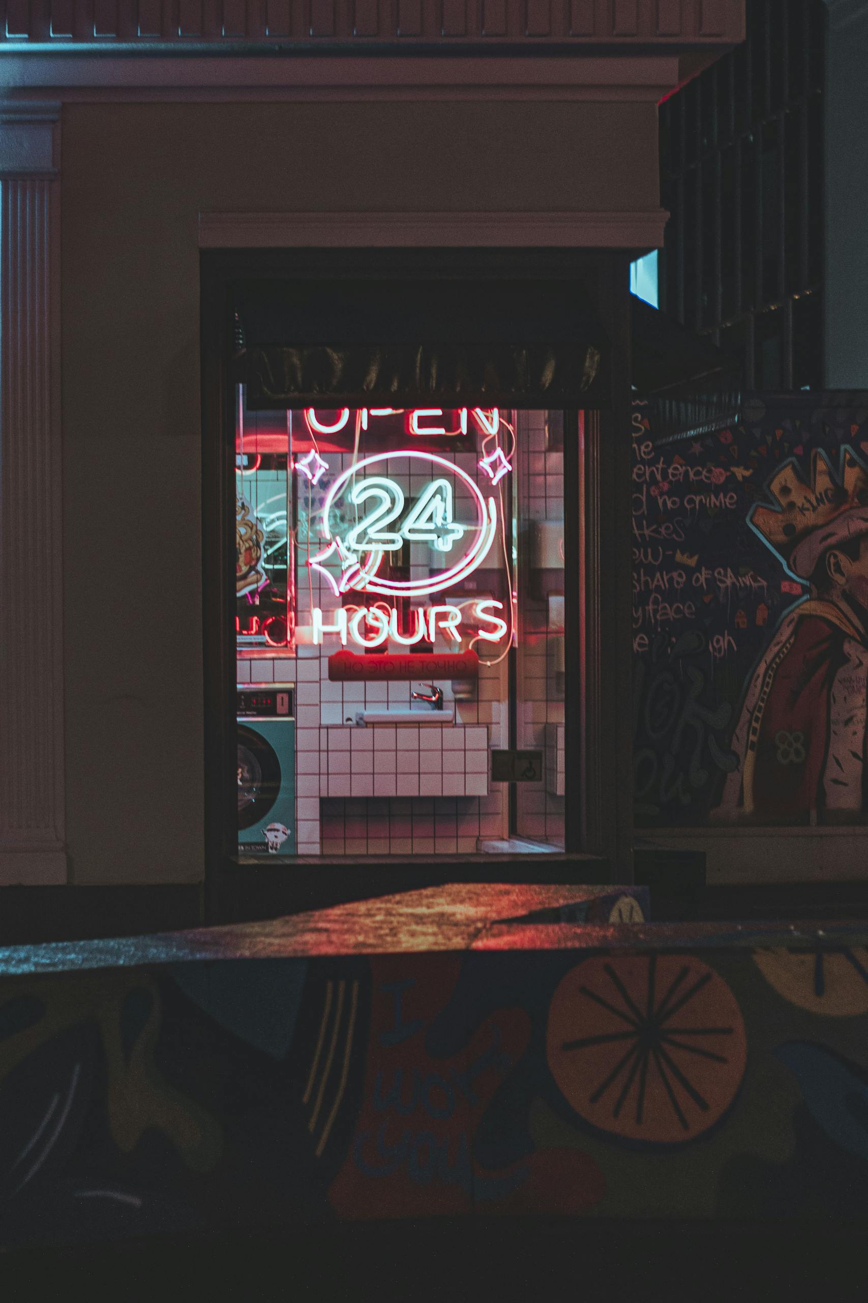 A vibrant neon "Open 24 Hours" sign illuminates an urban street with colorful graffiti art.